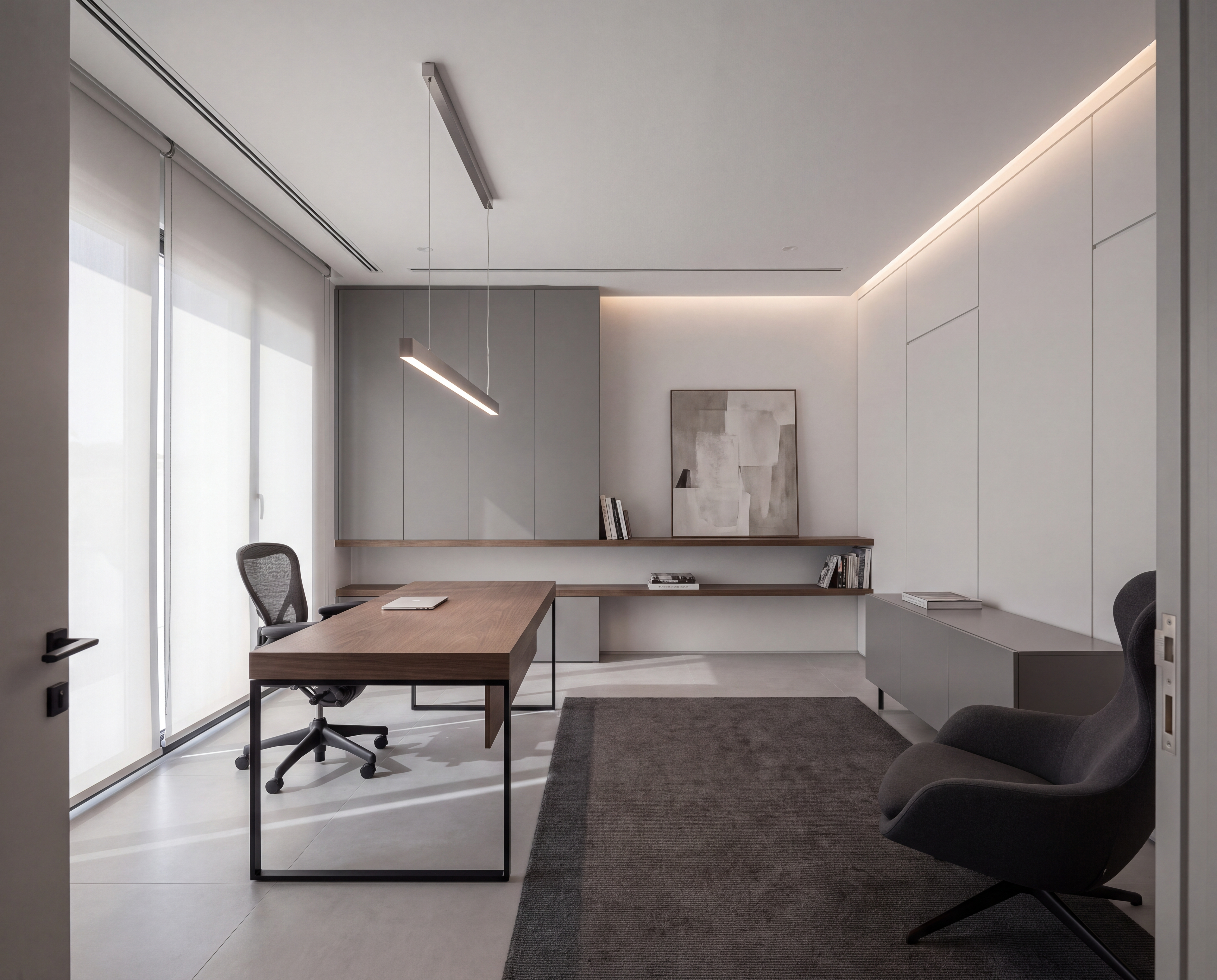 Modern home office with a smoked oak desk on a slim black frame, handleless grey cabinetry, a linear pendant light and a charcoal lounge chair