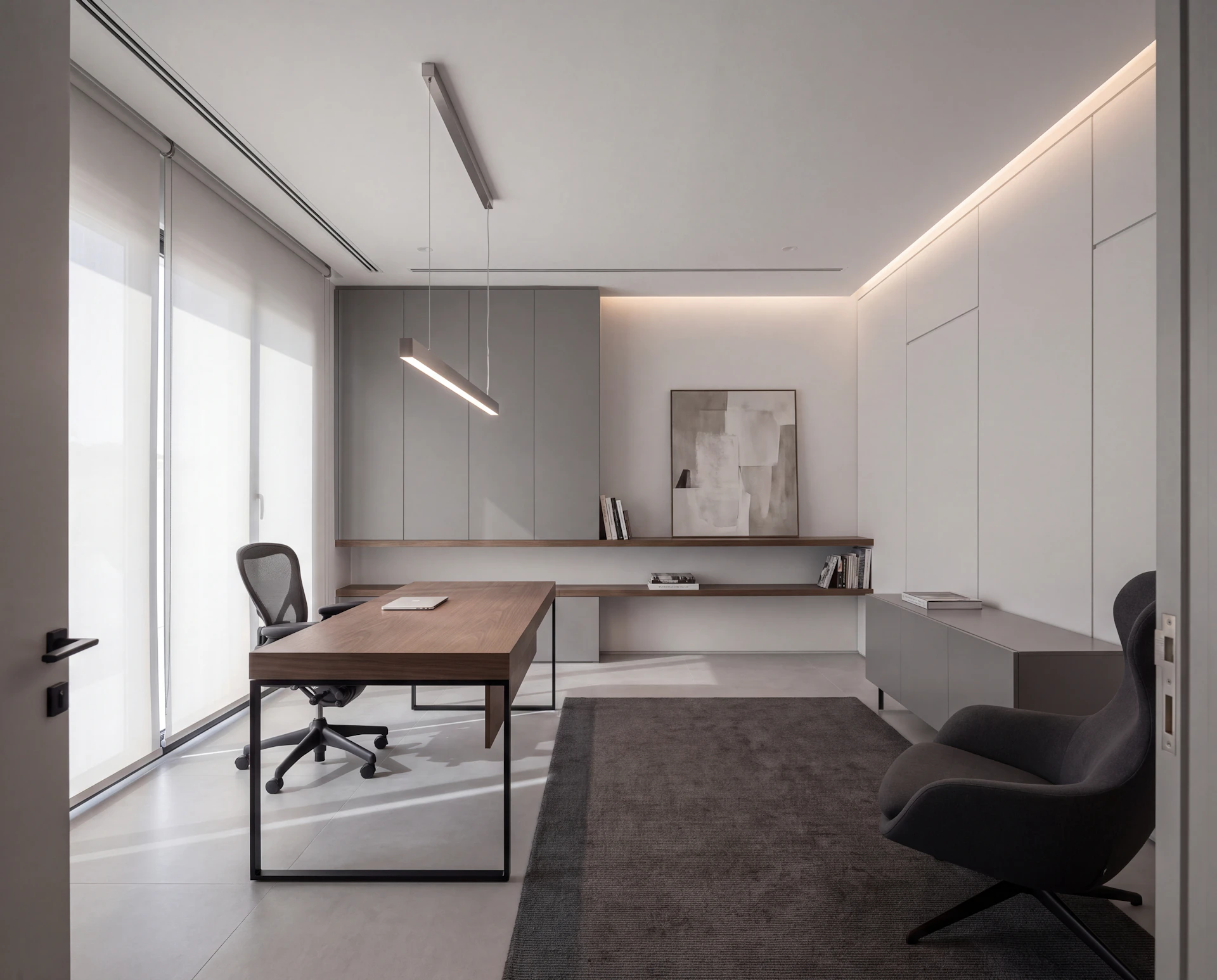 Modern home office with a smoked oak desk on a slim black frame, handleless grey cabinetry, a linear pendant light and a charcoal lounge chair